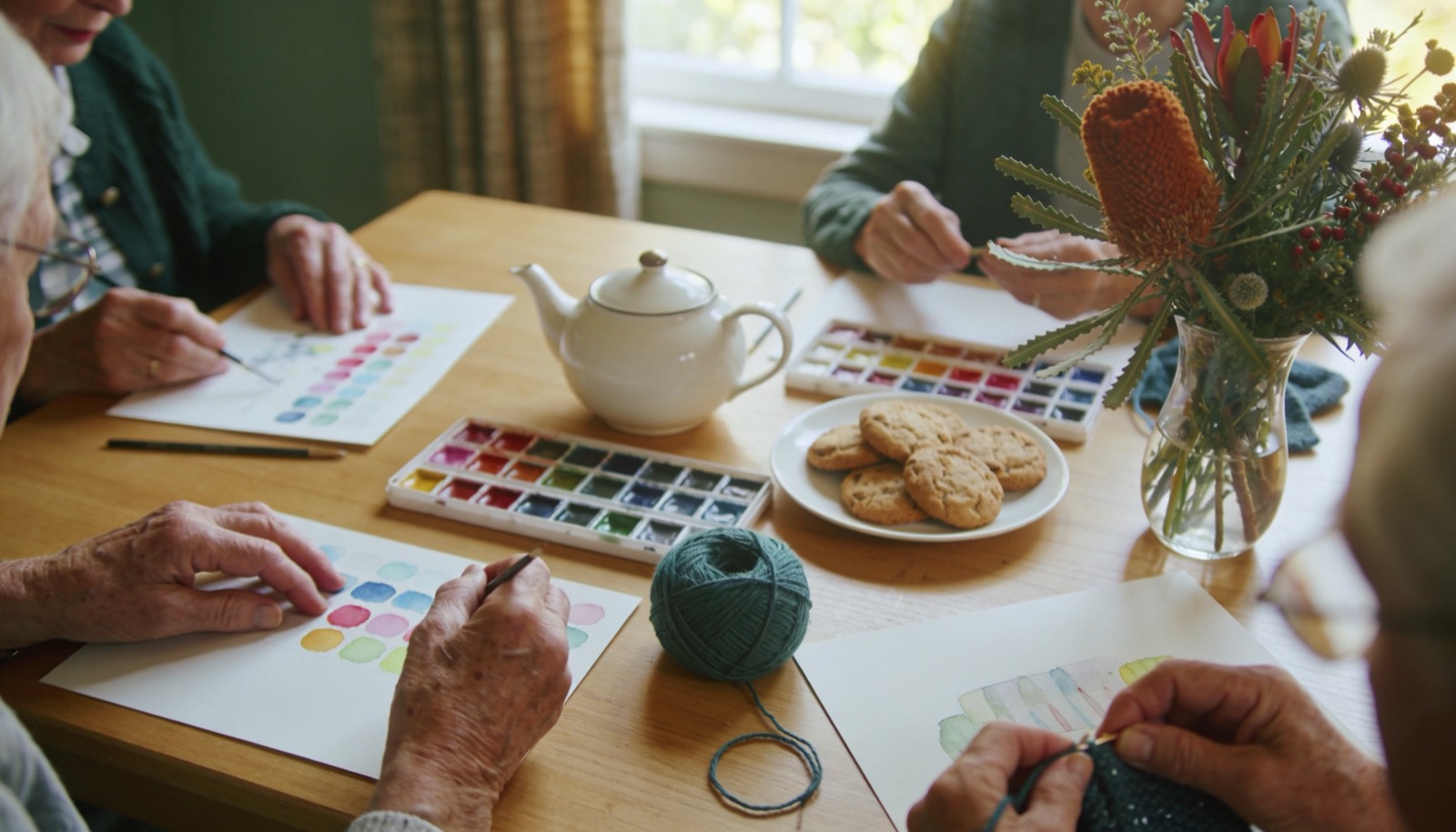 Hands at a sunlit table during a day-care art and craft session, with watercolours, knitting, a teapot, biscuits and native flowers.